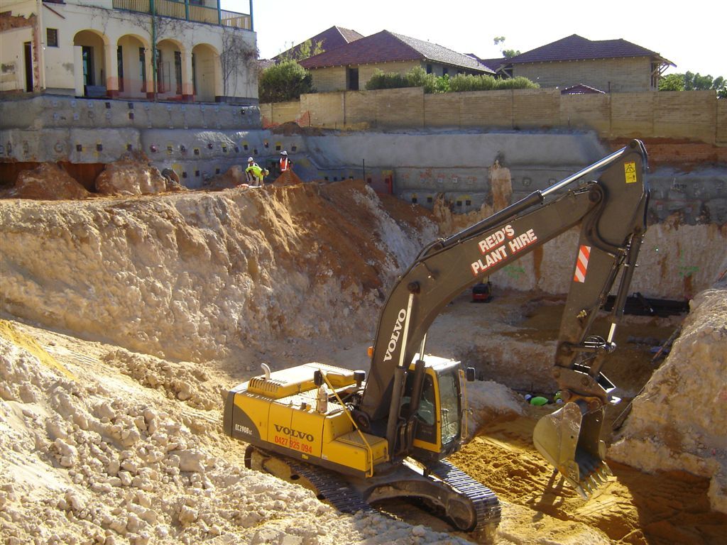Excavator at the bottom of a deep rock excavation beside an old brick building